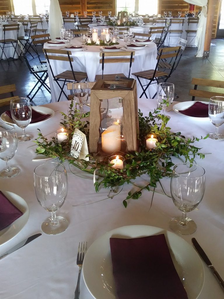 elegant rustic styled table setting with water goblets, white plates, burgundy folded napkins. The centerpiece is made with sprigs of eucalyptus, candles and a rustic lantern